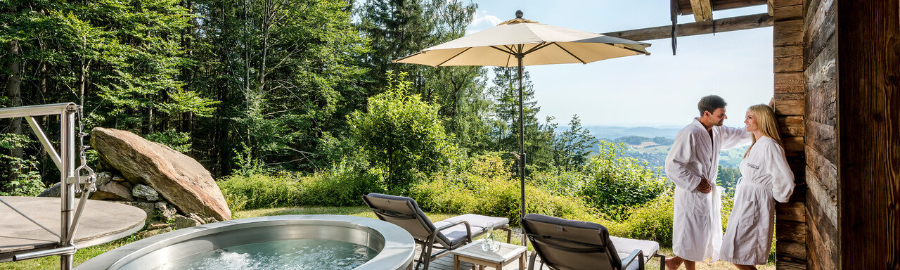 Pärchen im Bademantel steht auf Terrasse mit Whirlpool und Liegen des Luxus Chalets Bayerischer Wald