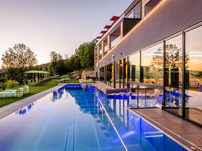 Blauer, beleuchteter Außenpool, umgeben von Liegen und Sonnenschirmen auf grüner Wiese