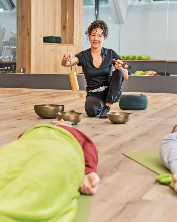Gäste liegen im Fitnessraum mit Spiegelwand auf Matten bei Meditationskurs um Dame mit Tonschalen