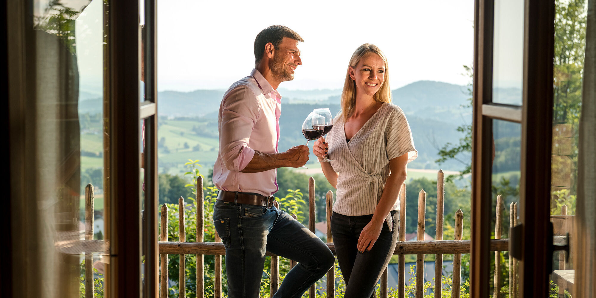 Luxus Chalet Bayern Terrasse mit Ausblick über das Bergdorf Hüttenhof und einem Pärchen das einen Wein genießt