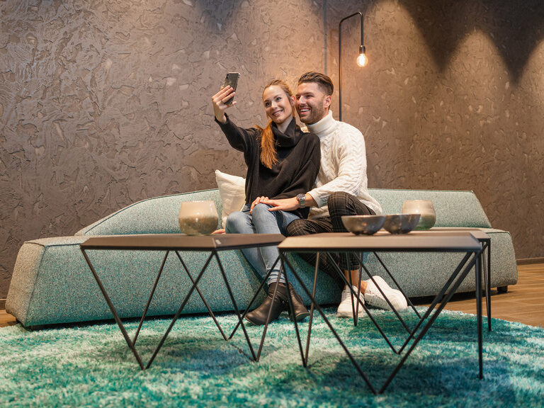 Paar sitzt auf Sofa in Loungebereich macht lachend ein Selfie mit dem Handy