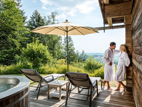 Pärchen im Bademantel steht auf Terrasse des Luxus Chalets Bayerischer Wald mit Whirlpool und Liegen