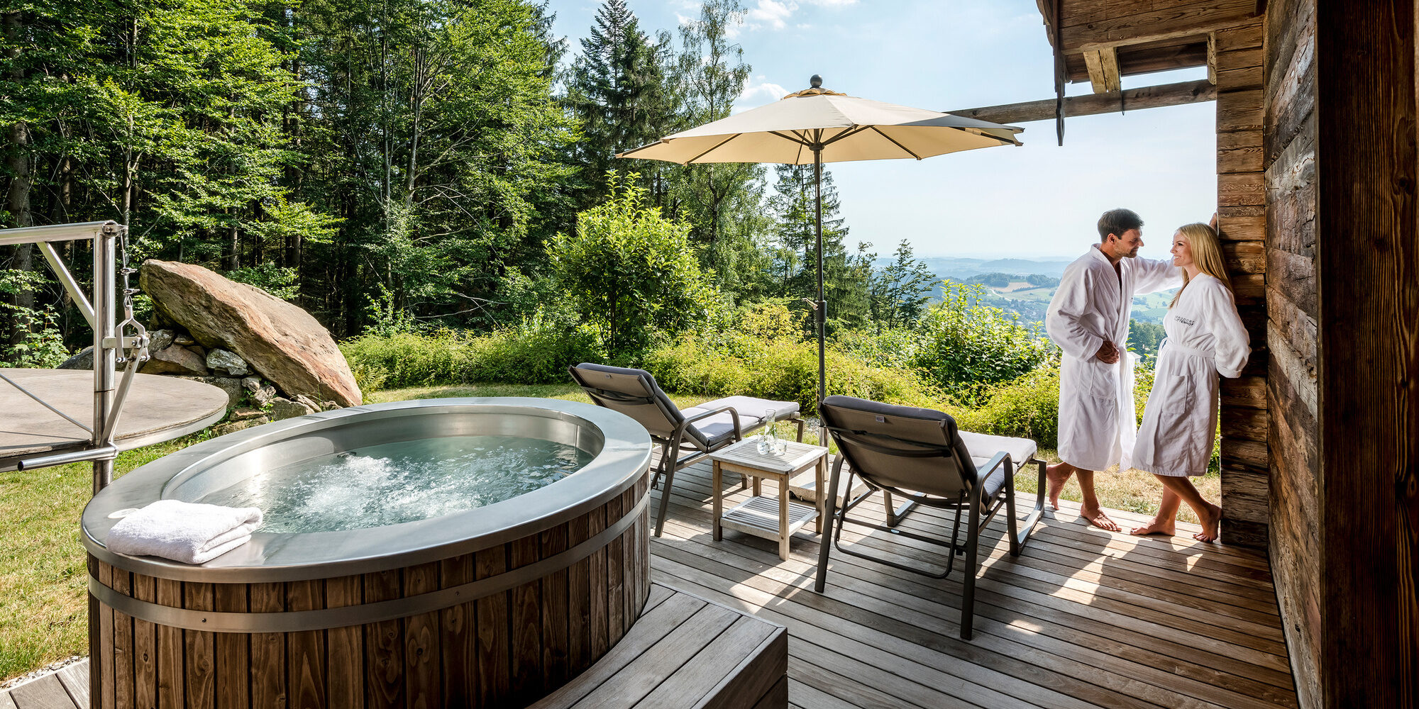 Pärchen im Bademantel steht auf Terrasse mit Whirlpool und Liegen des Luxus Chalets Bayerischer Wald